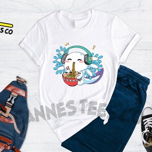 May include: White t-shirt with a cartoon axolotl wearing headphones and eating ramen noodles. The axolotl has blue and purple gills and is smiling. The text "Cannes Tees Co" is on the top left of the image.