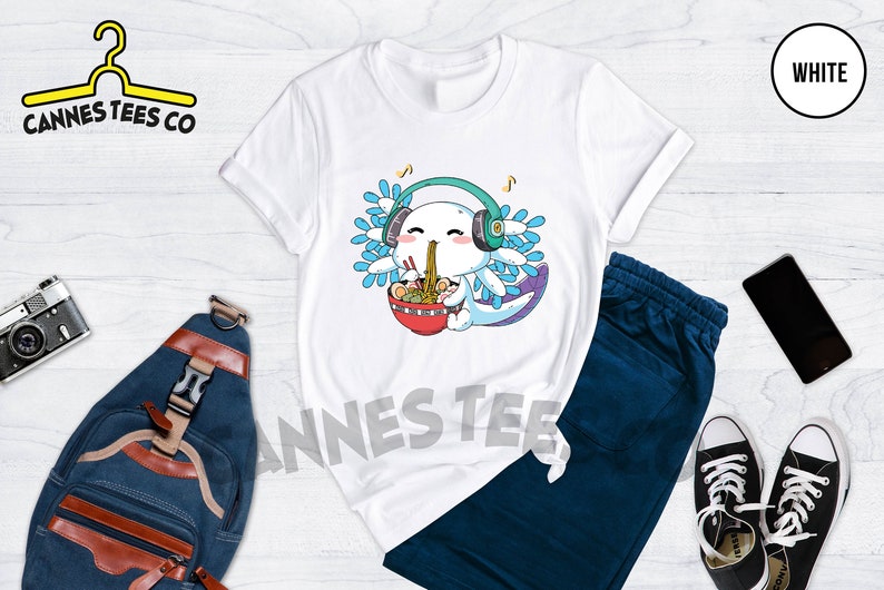 May include: White t-shirt with a cartoon axolotl wearing headphones and eating ramen noodles. The axolotl has blue and purple gills and is smiling. The text "Cannes Tees Co" is on the top left of the image.