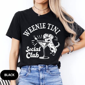 May include: Black t-shirt with white text that reads "Weenie Tini Social Club". A cartoon dachshund is holding a martini glass with an olive.