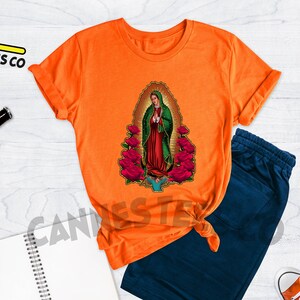 May include: Orange t-shirt with a graphic of Our Lady of Guadalupe surrounded by roses. The t-shirt is on a white wooden surface with a pair of blue sweatpants, a cell phone, and two pairs of red and white sneakers.
