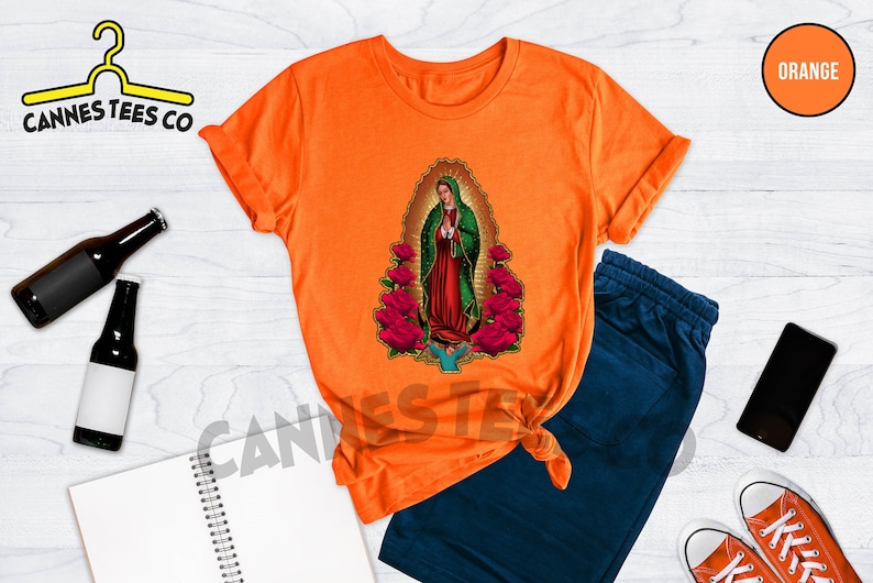 May include: Orange t-shirt with a graphic of Our Lady of Guadalupe surrounded by roses. The t-shirt is on a white wooden surface with a pair of blue sweatpants, a cell phone, and two pairs of red and white sneakers.