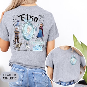 May include: Heather gray t-shirt featuring a Disney Frozen design. The back of the shirt displays the name "Elsa" above a framed portrait of Elsa, surrounded by other characters and the text "Disney Frozen Est. 2013". A smaller version of the design is on the front.