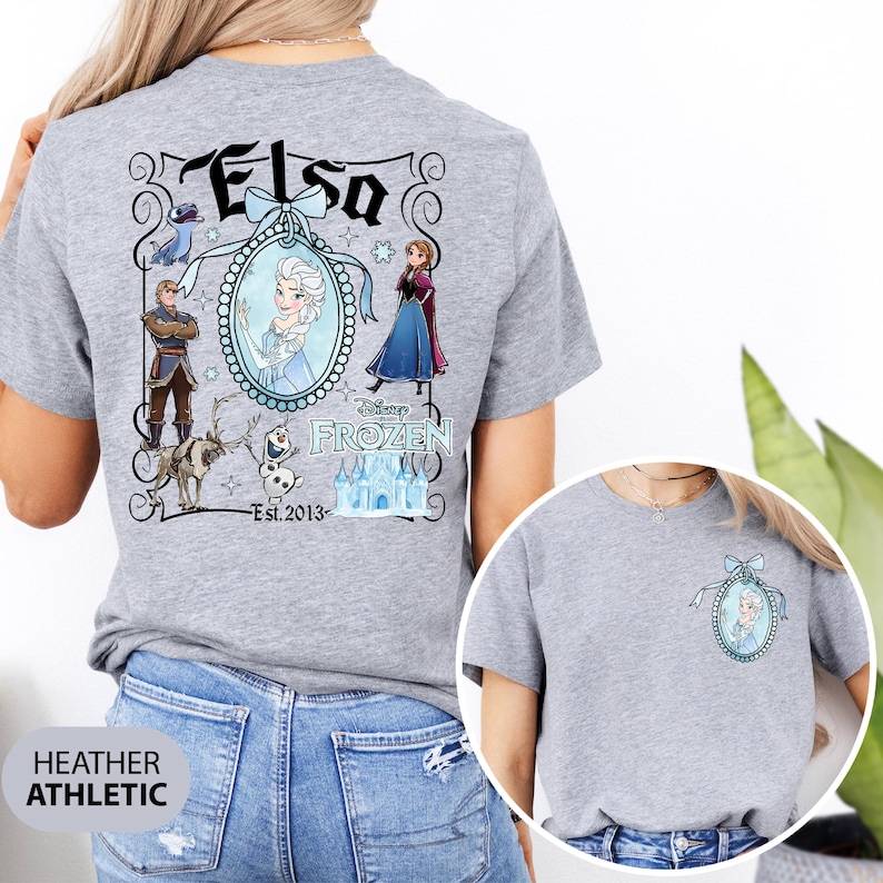 May include: Heather gray t-shirt featuring a Disney Frozen design. The back of the shirt displays the name "Elsa" above a framed portrait of Elsa, surrounded by other characters and the text "Disney Frozen Est. 2013". A smaller version of the design is on the front.