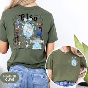 May include: Heather olive green t-shirt featuring a Disney Frozen design. The back of the shirt displays the name "Elsa" in a decorative frame with character illustrations. The front of the shirt has a smaller version of the Elsa design.