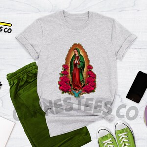 May include: A heather grey t-shirt with a graphic of Our Lady of Guadalupe surrounded by red roses. The t-shirt is laid flat on a wooden surface with other items, including a pair of green sweatpants, a notebook, a pair of green sneakers, a smartphone, and a skateboard.