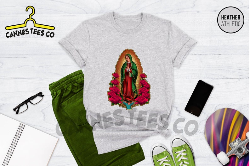May include: A heather grey t-shirt with a graphic of Our Lady of Guadalupe surrounded by red roses. The t-shirt is laid flat on a wooden surface with other items, including a pair of green sweatpants, a notebook, a pair of green sneakers, a smartphone, and a skateboard.