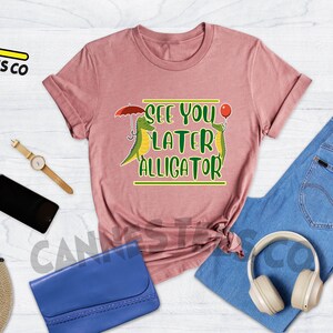 May include: Heather mauve t-shirt with a graphic of two green alligators, one holding an umbrella and the other holding a red balloon. The text "See you later alligator" is printed on the shirt.