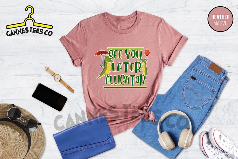 May include: Heather mauve t-shirt with a graphic of two green alligators, one holding an umbrella and the other holding a red balloon. The text "See you later alligator" is printed on the shirt.