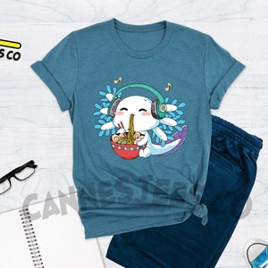 May include: A teal blue t-shirt with a cartoon axolotl wearing headphones and eating ramen noodles. The axolotl is surrounded by blue and white flowers. The text "Cannes Tees Co" is on the top left of the image.