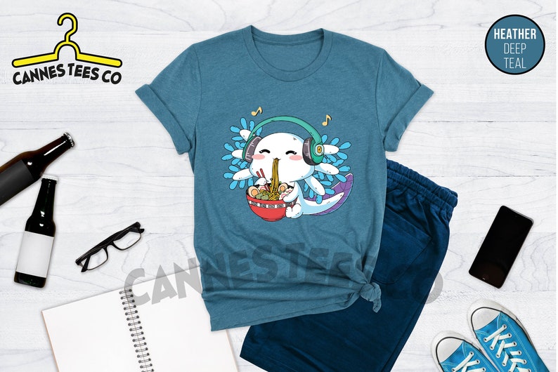 May include: A teal blue t-shirt with a cartoon axolotl wearing headphones and eating ramen noodles. The axolotl is surrounded by blue and white flowers. The text "Cannes Tees Co" is on the top left of the image.