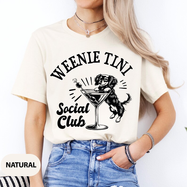 May include: A white t-shirt with a black graphic of a dachshund dog holding a martini glass. The text "Weenie Tini Social Club" is printed on the shirt.