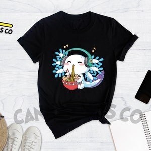 May include: Black t-shirt with a cartoon axolotl wearing headphones and eating ramen noodles. The axolotl is surrounded by blue and white flowers. The text "Cannes Tees Co" is on the top left of the image.