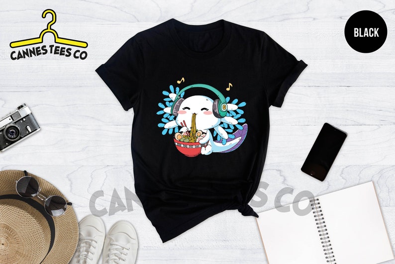 May include: Black t-shirt with a cartoon axolotl wearing headphones and eating ramen noodles. The axolotl is surrounded by blue and white flowers. The text "Cannes Tees Co" is on the top left of the image.