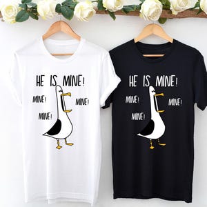 May include: Two t-shirts with a cartoon seagull graphic and the text "HE IS MINE!" repeated multiple times. The shirts are white and black.