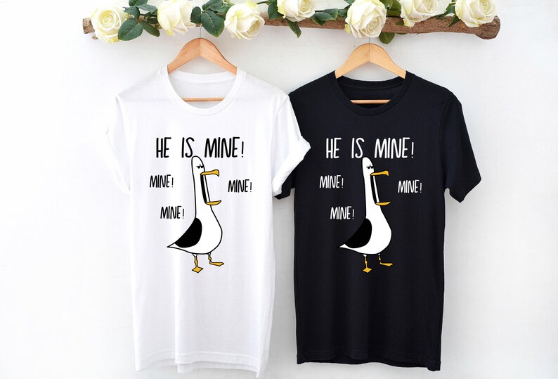 May include: Two t-shirts with a cartoon seagull graphic and the text "HE IS MINE!" repeated multiple times. The shirts are white and black.