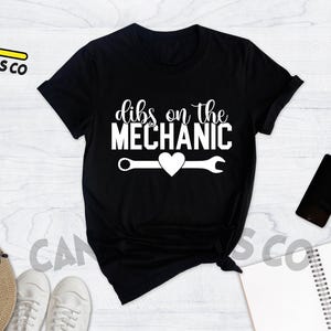 Może przedstawiać: Czarny T-shirt z białym napisem "Dibs on the Mechanic" z grafiką klucza i serca.