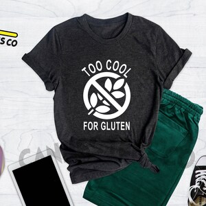 May include: A heather dark grey t-shirt with a white graphic of a crossed-out wheat stalk and the text "Too Cool For Gluten".