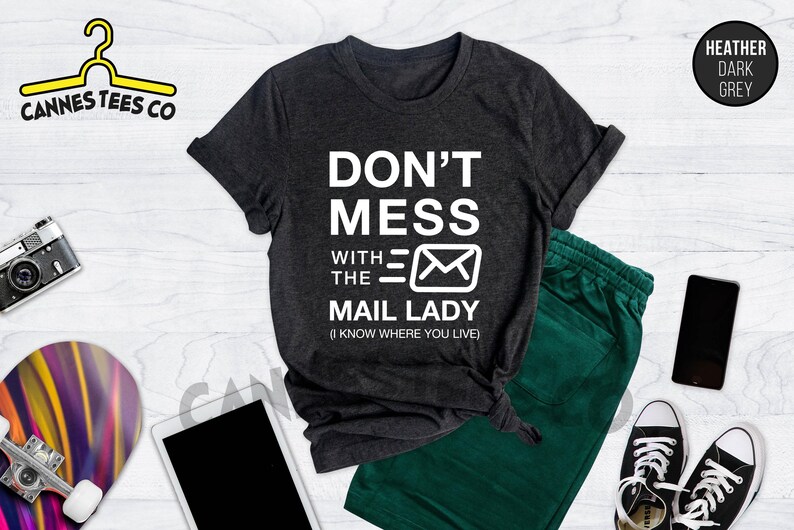 May include: A dark heather grey t-shirt with white text that reads "Don't Mess With The Mail Lady (I Know Where You Live)". The text is accompanied by a white envelope graphic.