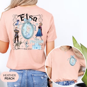 May include: Heather peach t-shirt featuring a Disney Frozen design. The back of the shirt displays the word "Elsa" above an oval portrait of Elsa, surrounded by other characters and the text "Est. 2013". A smaller version of the design is on the front.
