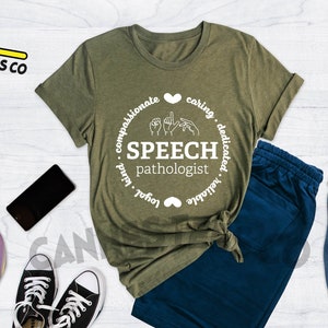 Op de afbeelding: Een groen T-shirt met een wit cirkelvormig ontwerp met de tekst "SPEECH pathologist" met gebarentaal voor het woord "speech". De cirkel bevat ook de woorden "compassionate, caring, dedicated, reliable, loyal, kind" rond de buitenkant.