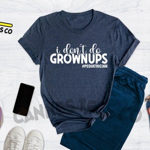May include: A heather navy blue t-shirt with the text "i don't do grown ups #pediatrician" in white. The shirt is on a white wooden surface with a pair of white sneakers, a skateboard, a camera, a phone, and a blue backpack.