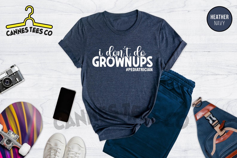 May include: A heather navy blue t-shirt with the text "i don't do grown ups #pediatrician" in white. The shirt is on a white wooden surface with a pair of white sneakers, a skateboard, a camera, a phone, and a blue backpack.