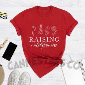 K&ouml;nnte beinhalten: Rotes T-Shirt mit wei&szlig;em Text "Raising Wildflowers" und einer Reihe von vier stilisierten Wildblumen.