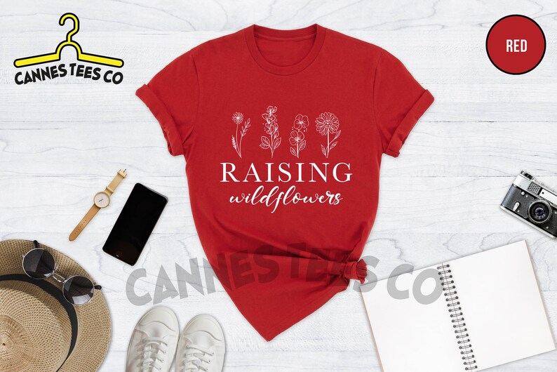 K&ouml;nnte beinhalten: Rotes T-Shirt mit wei&szlig;em Text "Raising Wildflowers" und einer Reihe von vier stilisierten Wildblumen.