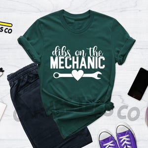 Może przedstawiać: Zielony T-shirt z białym napisem "dibs on the mechanic" i kluczem z sercem w środku.