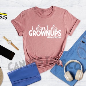 May include: A heather mauve t-shirt with the text "i don't do grown ups #pediatrician" printed in white. The shirt is laid out on a wooden surface with a pair of jeans, a hat, sunglasses, a phone, a watch, a purse, and headphones.
