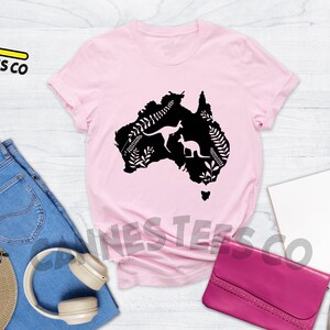 Może przedstawiać: R&oacute;żowy T-shirt z czarną sylwetką kontynentu australijskiego, przedstawiający dwa kangury i roślinność. Tekst "Cannes Tees Co" jest widoczny w lewym g&oacute;rnym rogu obrazu. Słowo "PINK" jest widoczne w prawym g&oacute;rnym rogu obrazu.
