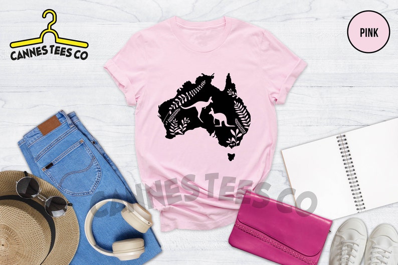 Może przedstawiać: R&oacute;żowy T-shirt z czarną sylwetką kontynentu australijskiego, przedstawiający dwa kangury i roślinność. Tekst "Cannes Tees Co" jest widoczny w lewym g&oacute;rnym rogu obrazu. Słowo "PINK" jest widoczne w prawym g&oacute;rnym rogu obrazu.