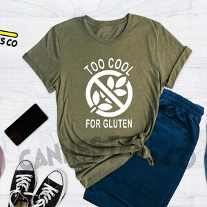 May include: A green t-shirt with the text "Too Cool For Gluten" in white. The text is inside a circle with a diagonal line through it and a wheat stalk graphic. The t-shirt is on a white wooden surface with a pair of blue jeans, a skateboard, a camera, a phone, and a backpack.