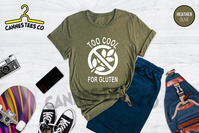 May include: A green t-shirt with the text "Too Cool For Gluten" in white. The text is inside a circle with a diagonal line through it and a wheat stalk graphic. The t-shirt is on a white wooden surface with a pair of blue jeans, a skateboard, a camera, a phone, and a backpack.