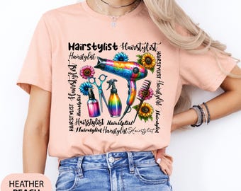 May include: A pink t-shirt with a colorful graphic design featuring a hairdryer, scissors, and hairspray bottles. The graphic also includes the text "Hairstylist" repeated multiple times.
