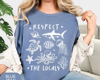 May include: Blue jean colored t-shirt with a white graphic design featuring various sea creatures and the text "RESPECT THE LOCALS". The design includes a seashell, shark, turtle, fish, jellyfish, starfish, octopus, and coral.