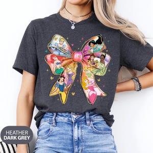 May include: Heather dark grey t-shirt featuring a large, colourful bow with images of Disney princesses. The bow is made of gold glitter and has a sparkly effect.