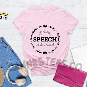 Op de afbeelding: Roze T-shirt met een zwart cirkelvormig ontwerp met de tekst "Speech pathologist" met handgebaren voor het woord "speech" en de woorden "compassionate, caring, dedicated, valuable, loyal, kind" rond de cirkel.