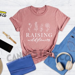 K&ouml;nnte beinhalten: Ein T-Shirt in Heide-Mauve mit dem Text "Raising wildflowers" und einer Strichzeichnung von vier Wildblumen. Das Shirt liegt auf einer wei&szlig;en Holzoberfl&auml;che mit einer blauen Jeans, einem Strohhut, einer Sonnenbrille, einer Uhr, einer blauen Clutch und Kopfh&ouml;rern.