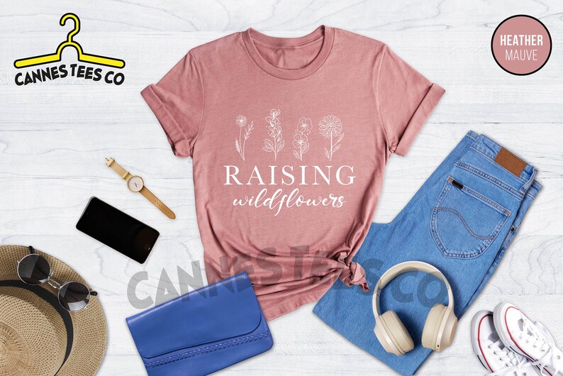 K&ouml;nnte beinhalten: Ein T-Shirt in Heide-Mauve mit dem Text "Raising wildflowers" und einer Strichzeichnung von vier Wildblumen. Das Shirt liegt auf einer wei&szlig;en Holzoberfl&auml;che mit einer blauen Jeans, einem Strohhut, einer Sonnenbrille, einer Uhr, einer blauen Clutch und Kopfh&ouml;rern.