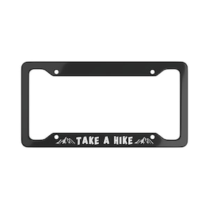Puede incluir: Marco de matrícula negro con el texto "TAKE A HIKE" en blanco, con un gráfico de cordillera. El marco tiene forma rectangular con esquinas redondeadas y cuatro orificios de montaje.