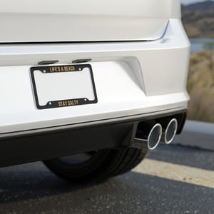 May include: A white car with a black license plate frame that says "Life's a Beach" and "Stay Salty" in gold lettering. The car has dual exhaust pipes.