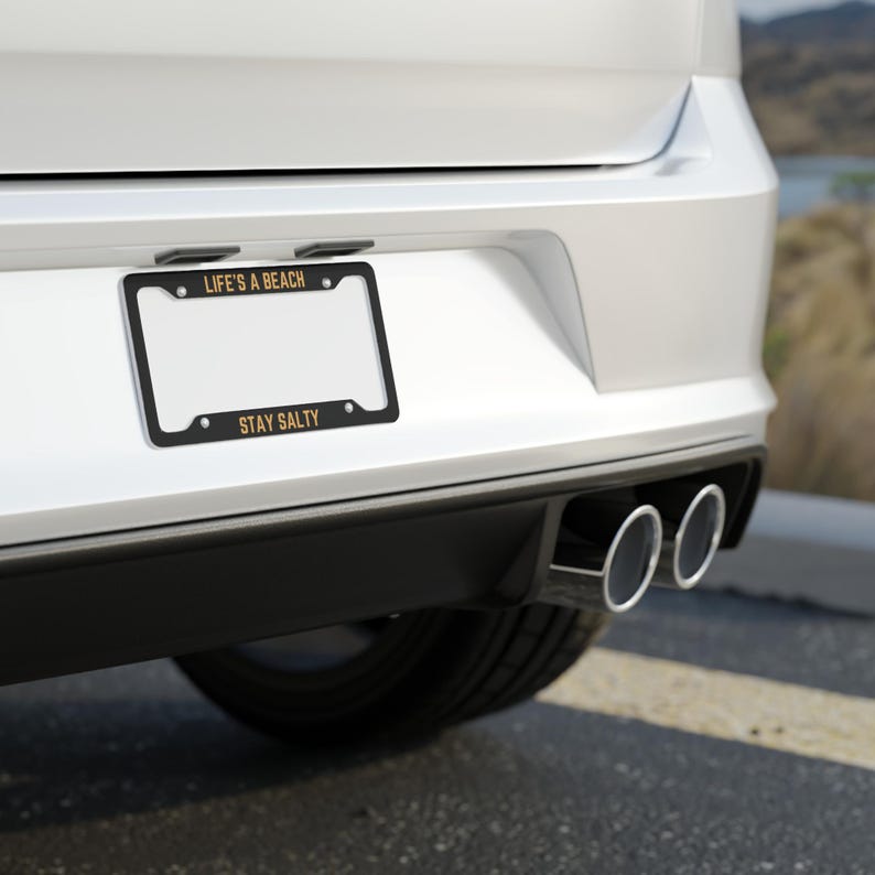 May include: A white car with a black license plate frame that says "Life's a Beach" and "Stay Salty" in gold lettering. The car has dual exhaust pipes.