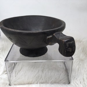 May include: A dark brown wooden bowl with a carved handle shaped like a face. The bowl has a wide, shallow base and a rounded rim.