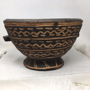 May include: A dark brown wooden bowl with a carved geometric design in a lighter brown colour. The bowl has two small handles on the sides and a wide base.
