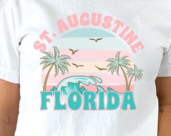 Może przedstawiać: Biała koszulka z nadrukiem graficznym przedstawiającym zachód słońca nad palmami i falami oceanu. Tekst "St. Augustine Florida" jest wydrukowany retro czcionką.