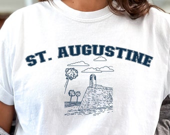 Peut inclure: T-shirt blanc avec un graphique bleu d'un phare et de palmiers. Le texte "ST. AUGUSTINE" est imprimé en bleu au-dessus du graphique.