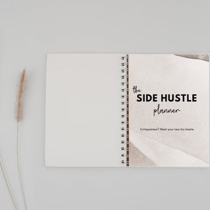 May include: A closed spiral bound notebook with a white cover and a brown and white abstract design on the front. The text "the SIDE HUSTLE planner" is printed in black on the front cover. The text "Entrepreneur? Meet your new biz bestie." is printed in black below the title.