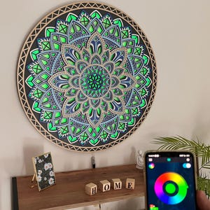 Könnte beinhalten: Ein großes, rundes Wandkunstwerk aus Holz mit Mandala-Motiv und mehreren Schichten, mit einem komplexen geometrischen Design in Grün, Blau und Schwarz. Das Mandala wird mit LED-Leuchten beleuchtet. Ein Smartphone zeigt ein Farbrad zur Steuerung der Lichter.