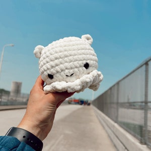 May include: A white crocheted plush toy with black eyes and a small smile. The toy is being held in a hand against a blue sky background.